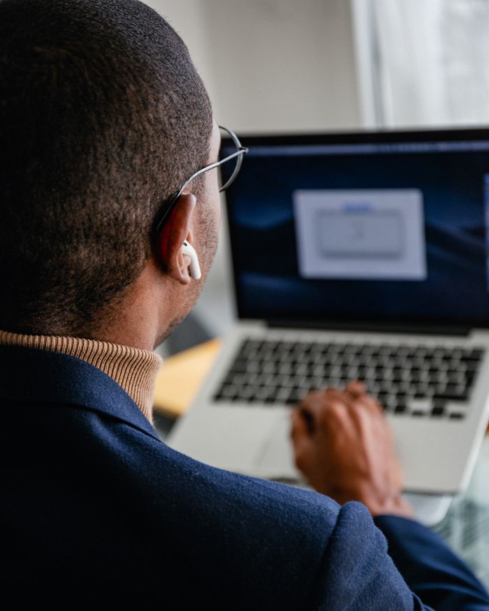 Back view of a man using a laptop with earbuds, focusing on his task indoors.
